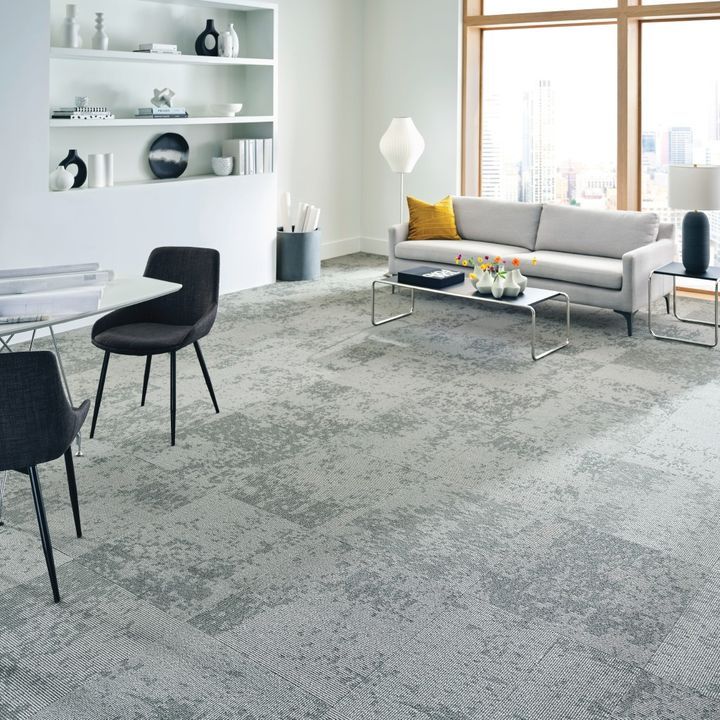 Modern office lounge with gray patterned carpet, sofa, table, chairs, and large window.