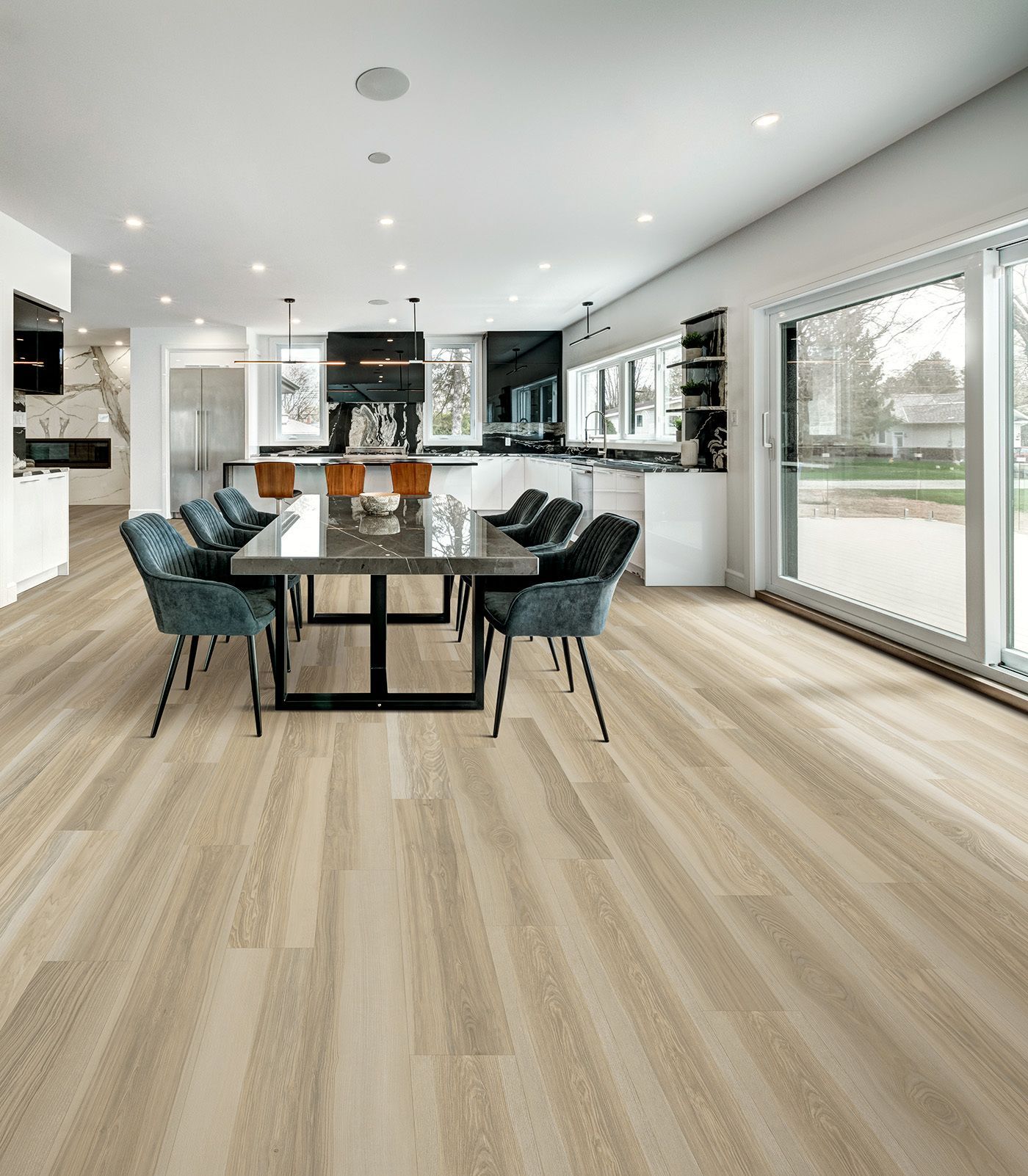 Spacious modern dining area with a long table, blue chairs, and wood-look flooring. White kitchen in the background.