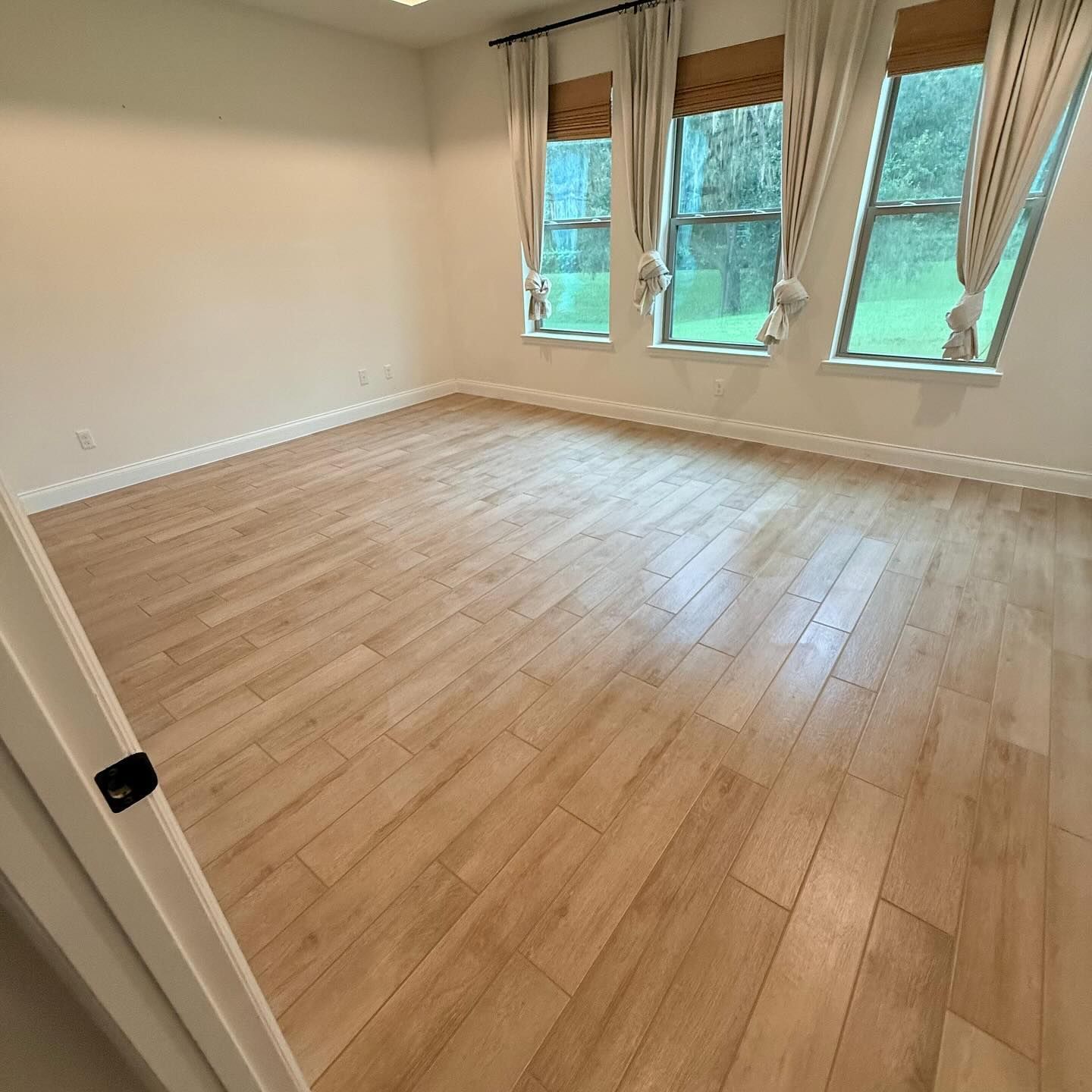 Empty room with wood-look floor, three windows with curtains, and a door on the left.