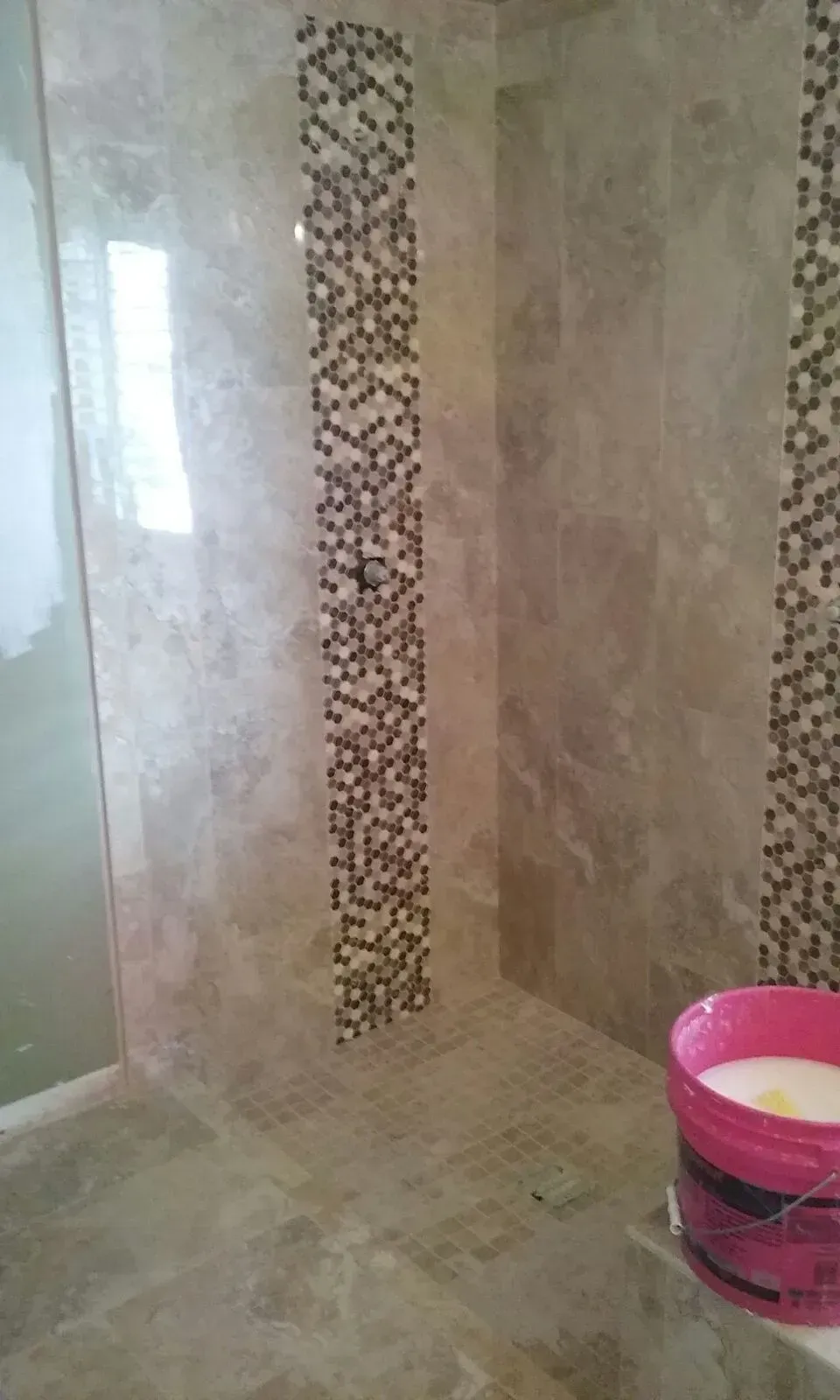 Shower stall with beige tile, mosaic accent strip, and a pink bucket of adhesive.