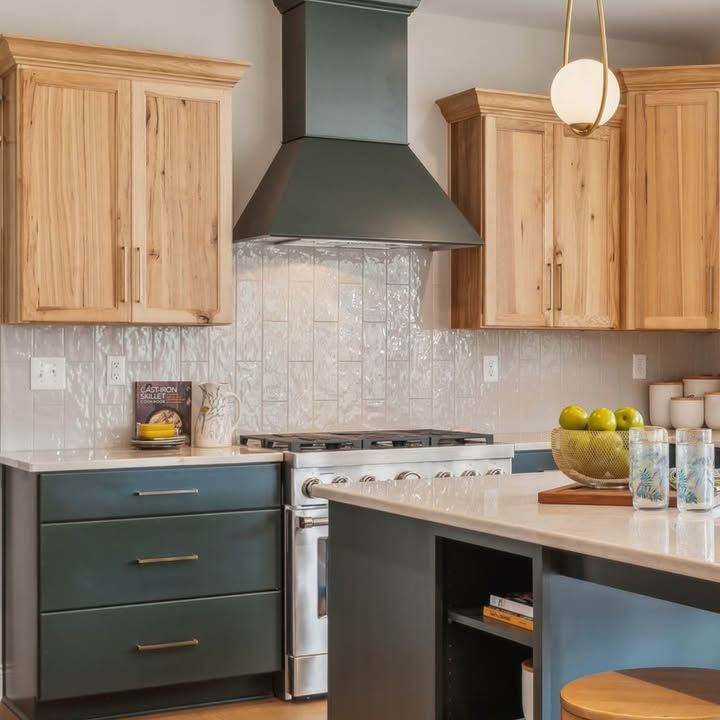 Kitchen with natural wood upper cabinets, teal lower cabinets, a dark range hood, and white tile backsplash.