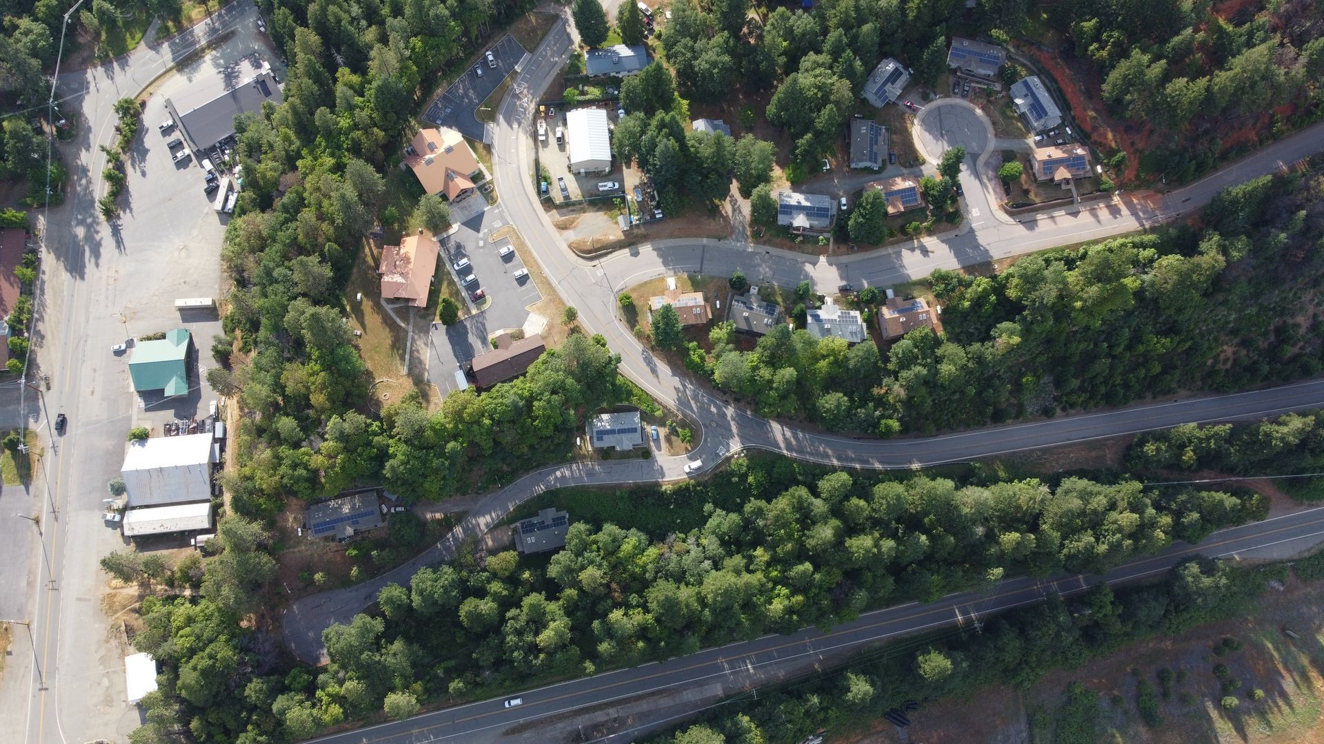 Aerial view of a town surrounded by trees and winding roads. Buildings and parking areas are visible.