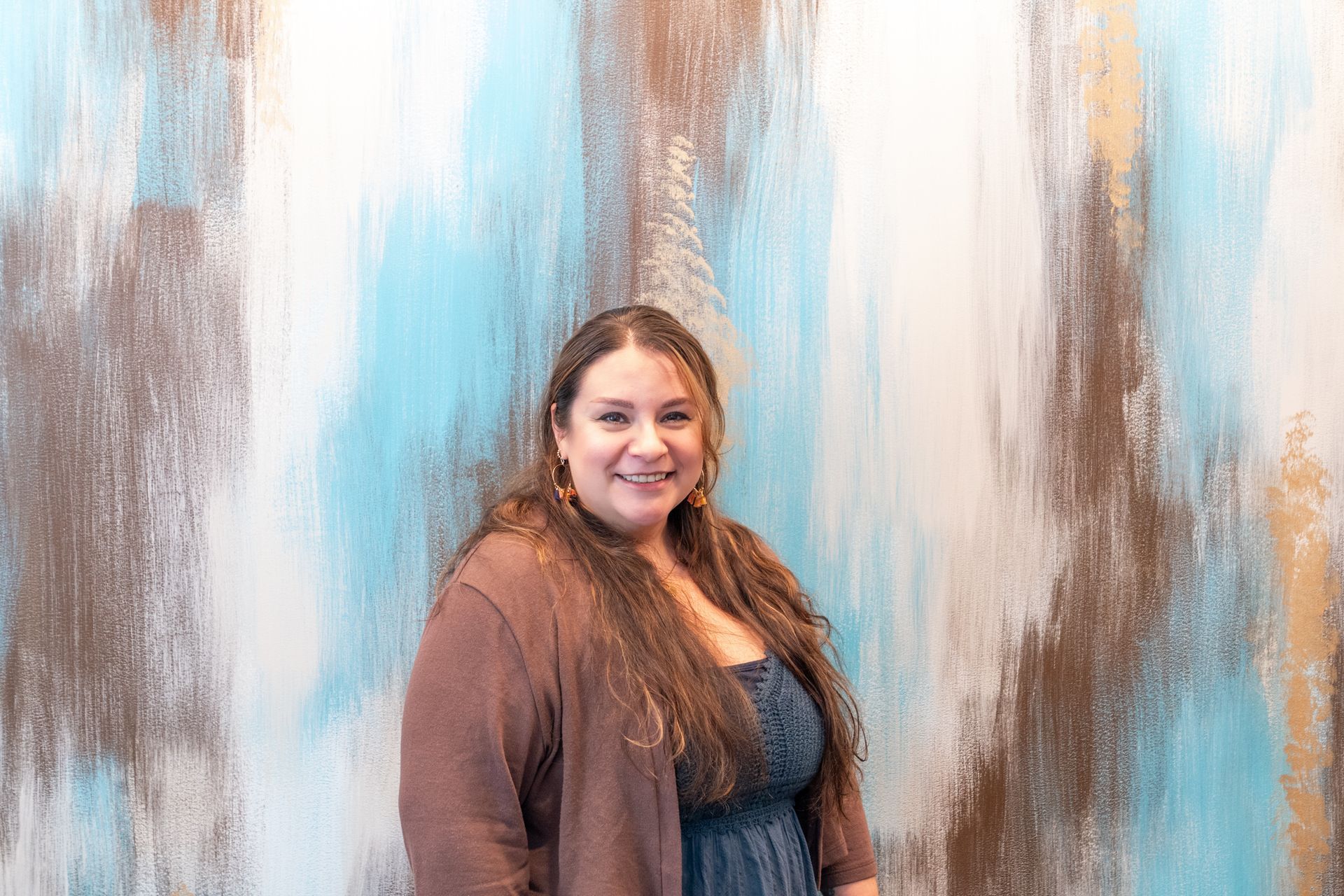Woman with long brown hair smiling in front of an abstract blue, brown, and white wall.