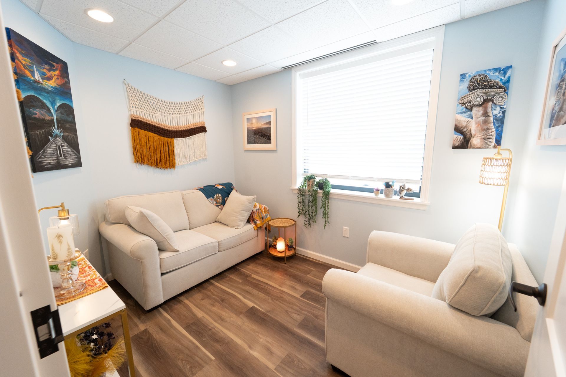 Cozy room with cream sofa, armchair, and art on light blue walls, wood floor, window with blinds.