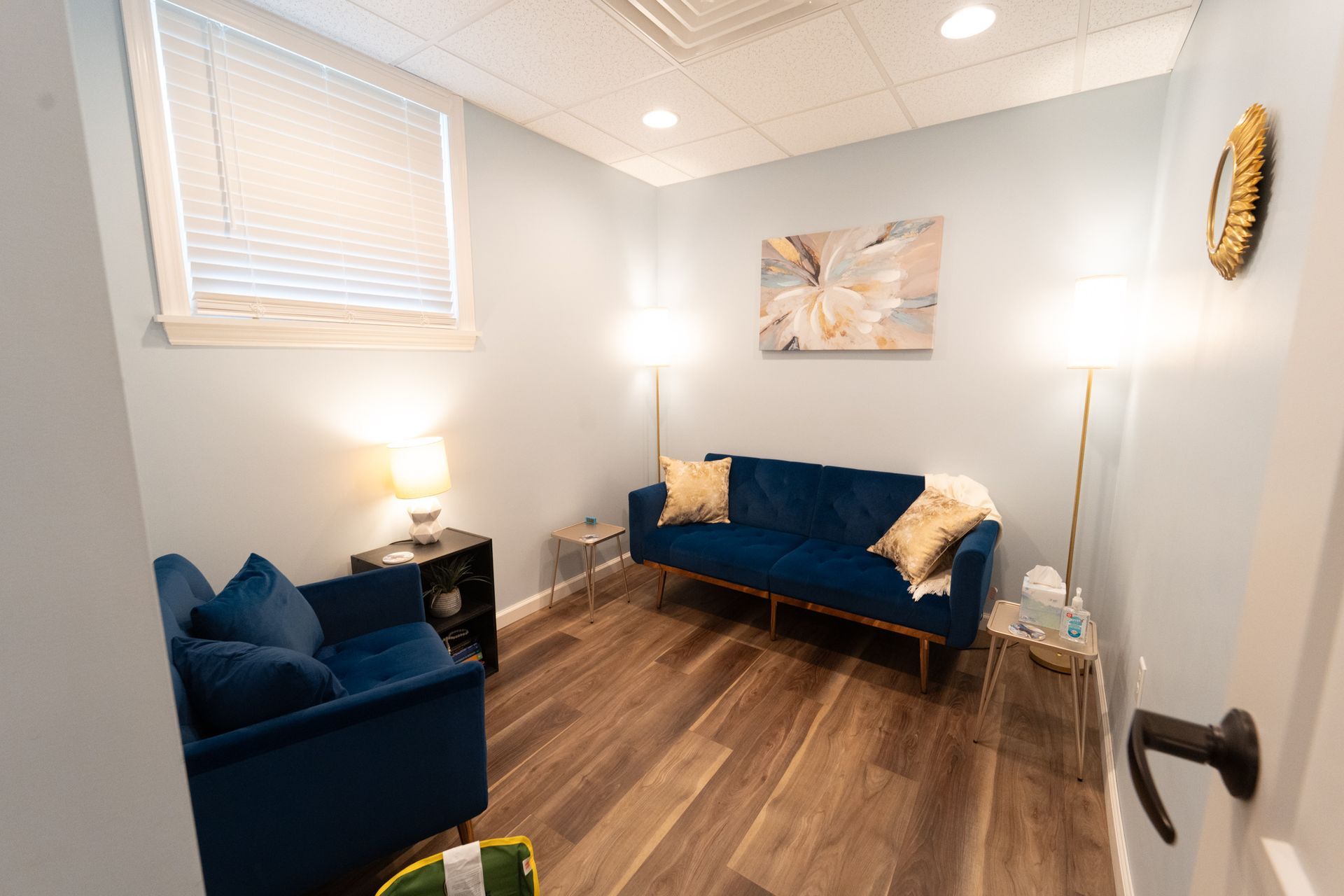 Therapy room: blue velvet sofa and chair, hardwood floors, art, lamps, and a window.