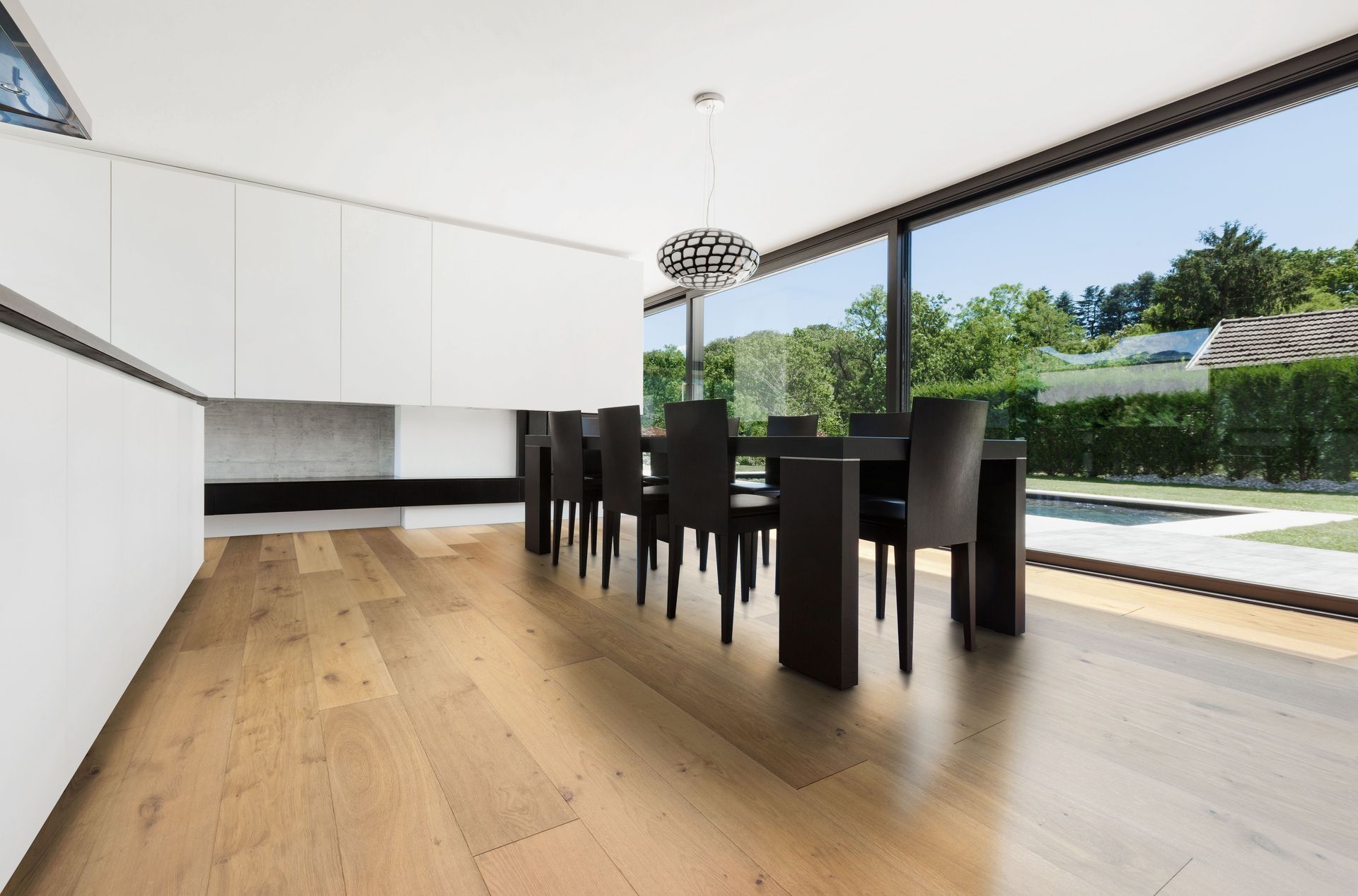 Modern dining room with a long black table and chairs, light wood floor, and large window overlooking a yard.