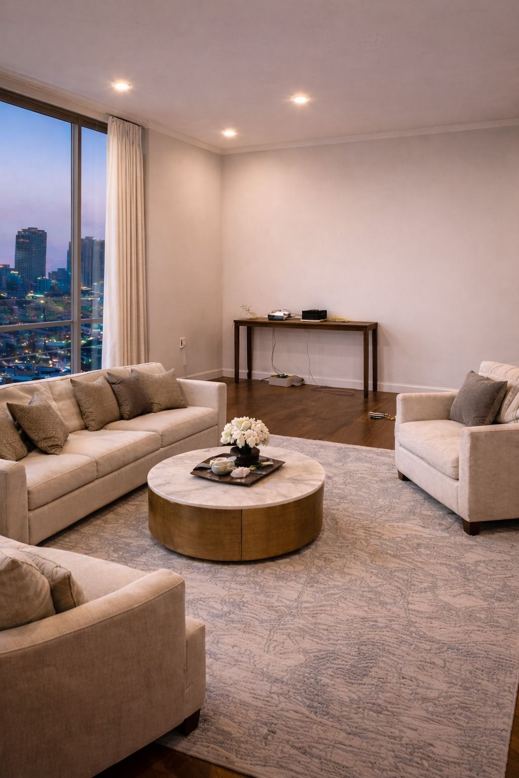 A modern living room with cream sofas, a round gold coffee table, and a console table, overlooking a city skyline.