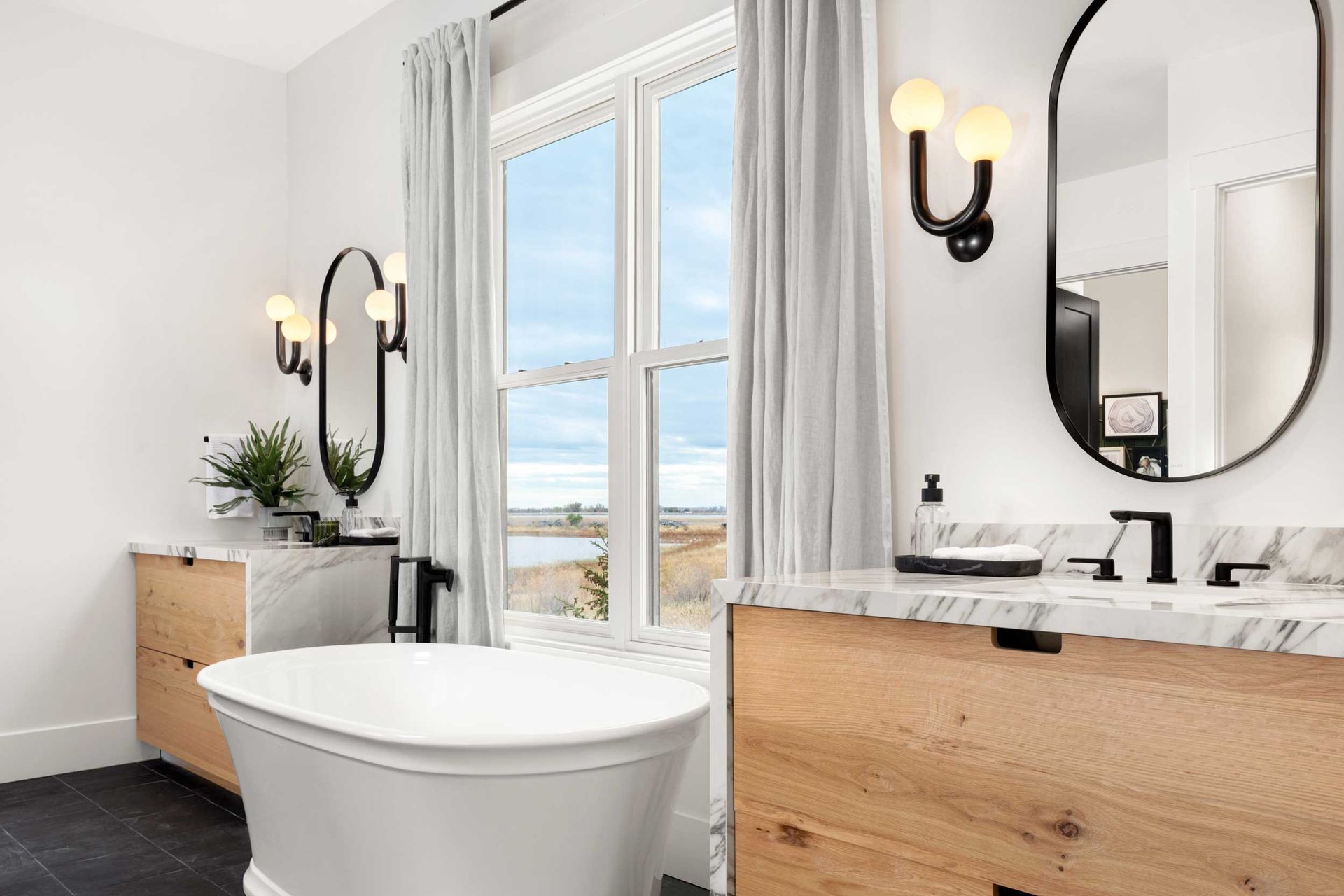 Modern bathroom with a bathtub, two sinks, mirrors, and a window overlooking an outdoor view.