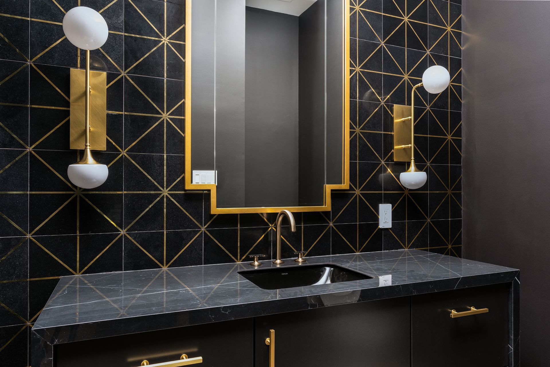 Black and gold bathroom with geometric tile, gold mirror, sink, and sconces.