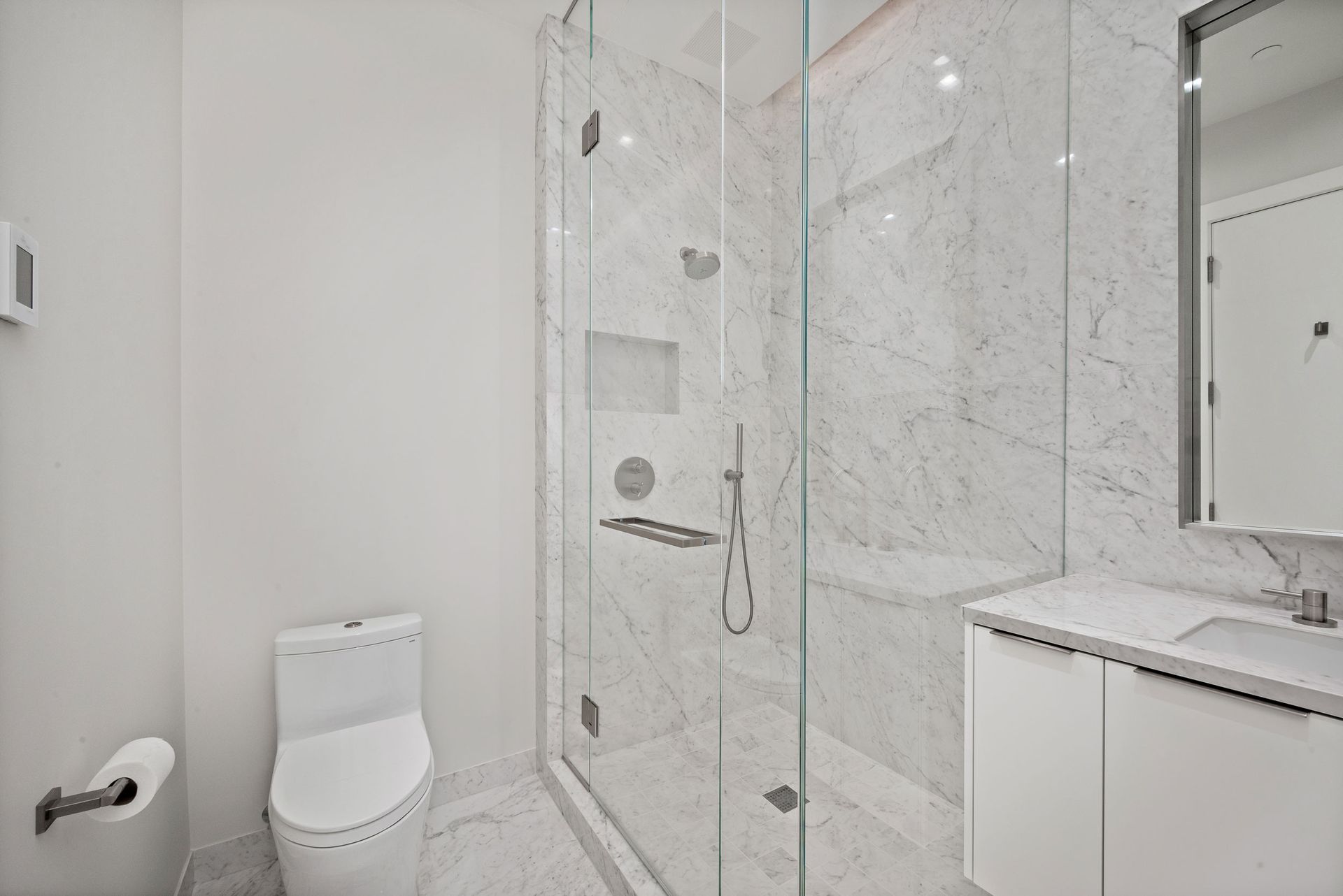 Bathroom with white marble walls, glass shower, white toilet, and floating vanity.