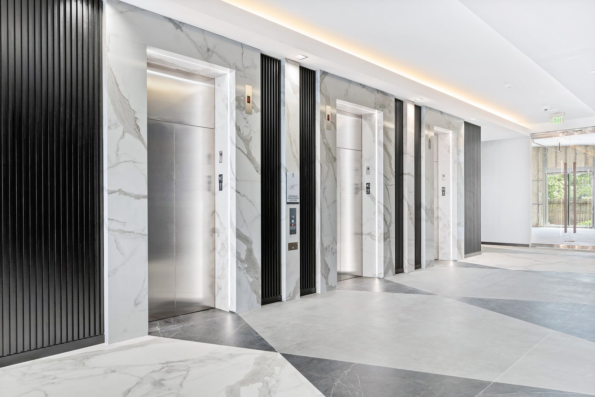 Modern elevator lobby with three stainless steel elevators, marble walls, and geometric tile floor.