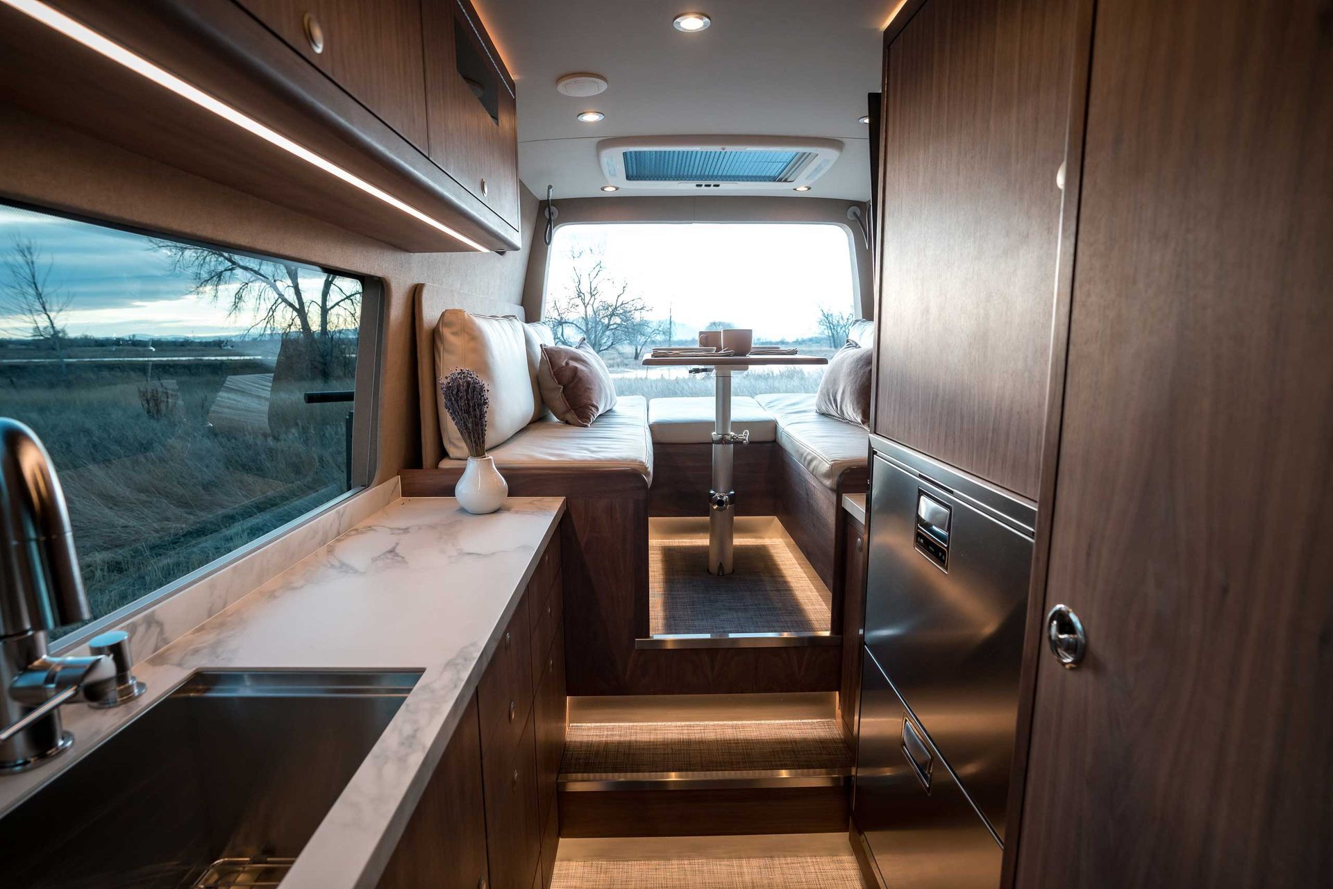 Interior of a modern camper van with wooden cabinets, marble countertop, and seating area near a window.