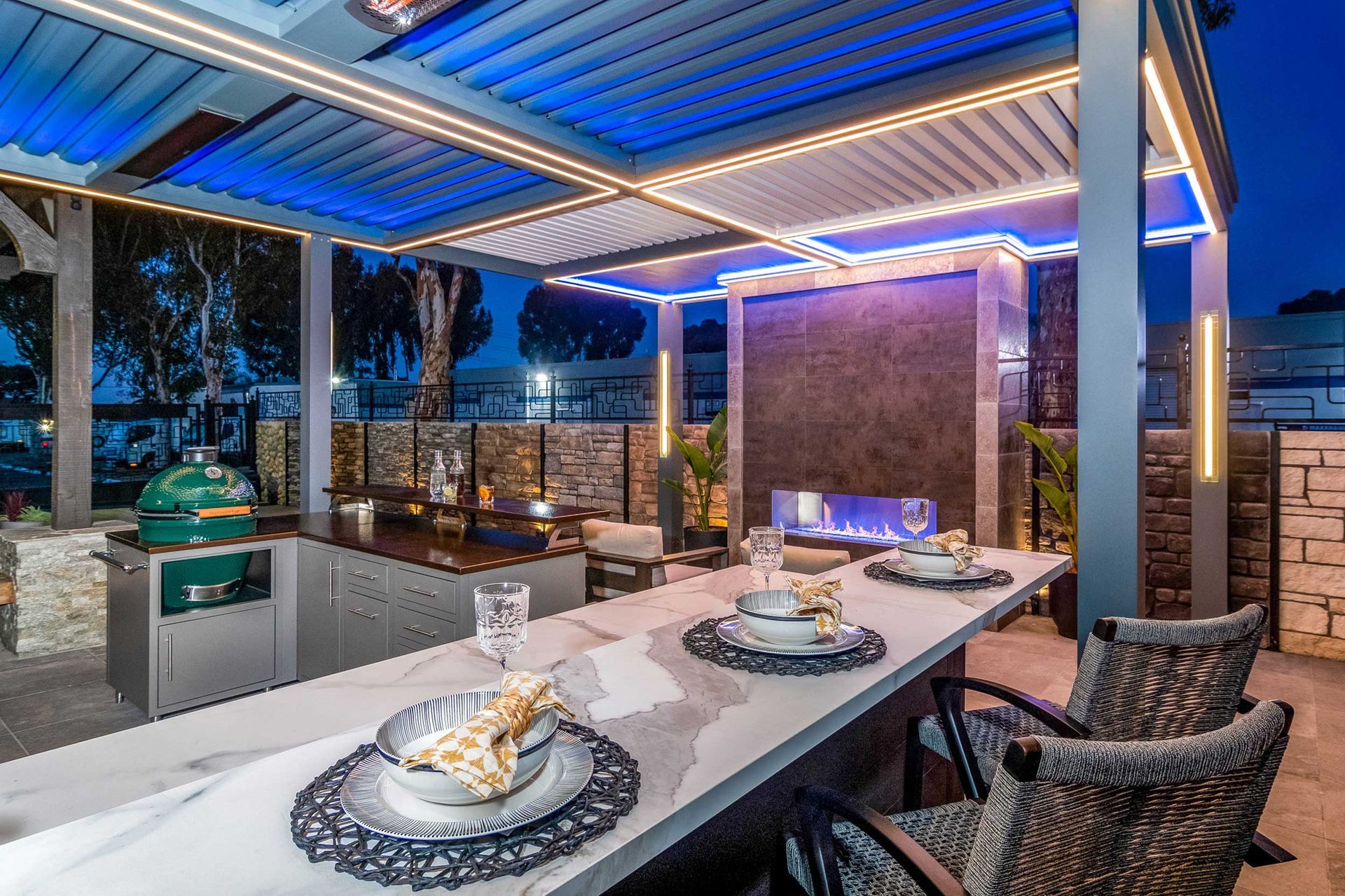 Outdoor bar with grill, fireplace, and overhead blue lighting. Marble countertop, wicker seating, and stone accents.