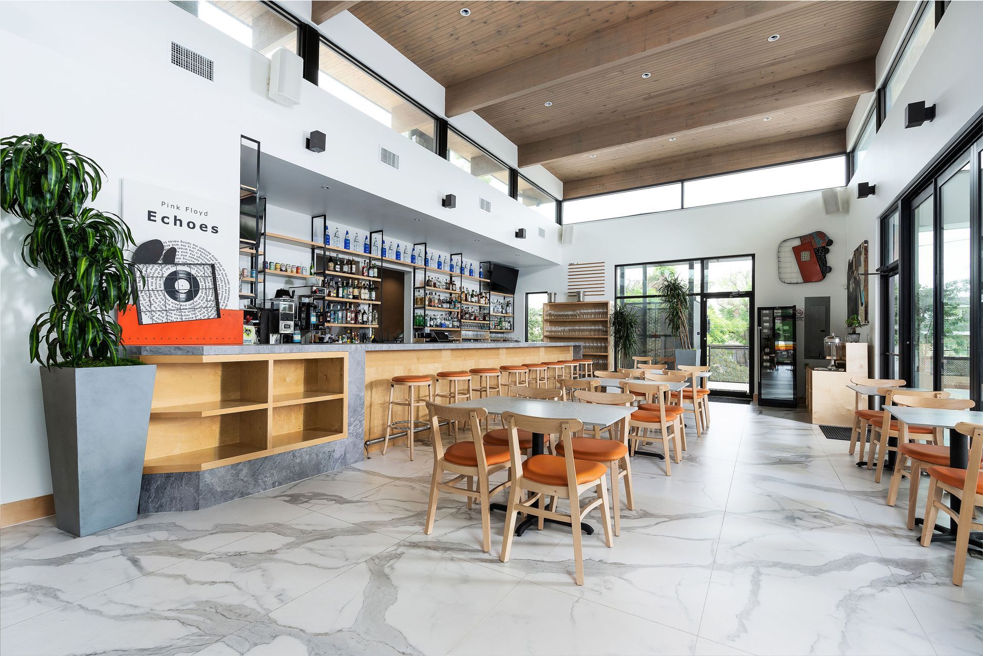 Interior of a modern cafe with bar, tables, chairs, plants, and large windows. Bright space with marble floors.