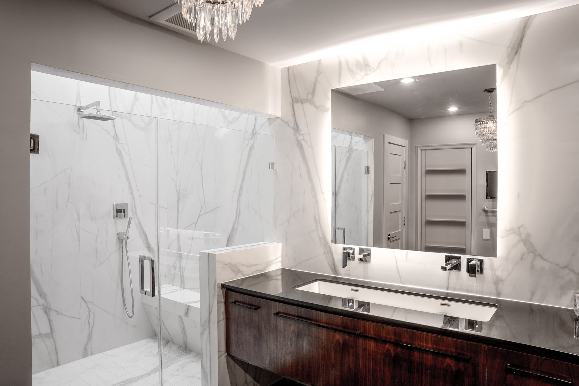 Luxurious white marble bathroom with a large shower, vanity, and crystal chandelier.