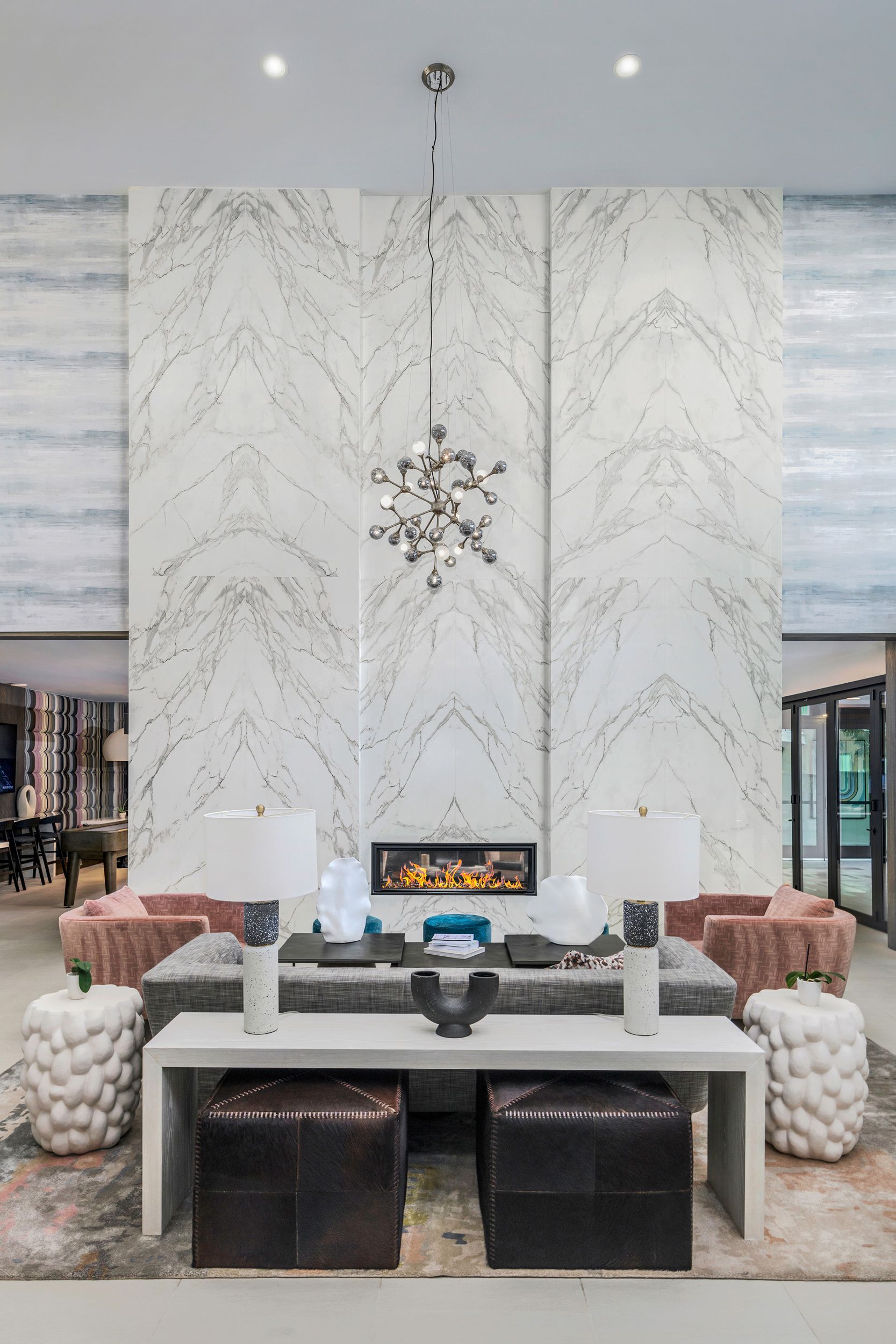 Lobby with a marble-like patterned wall, fireplace, modern chandelier, seating, and decorative ottomans.