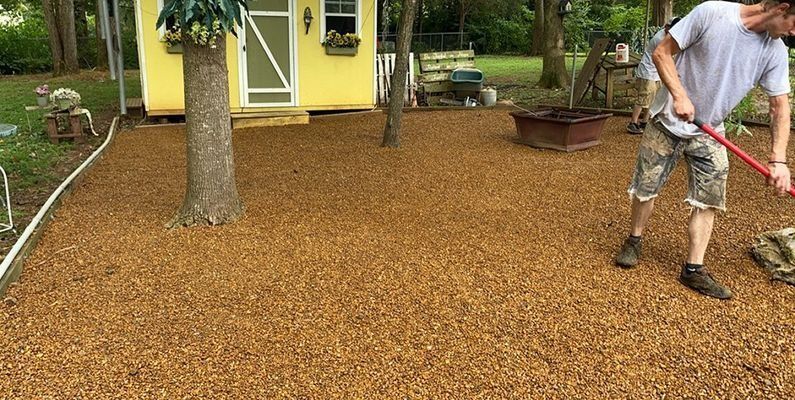 A person rakes mulch near a yellow playhouse, trees, and a fire pit in a yard.