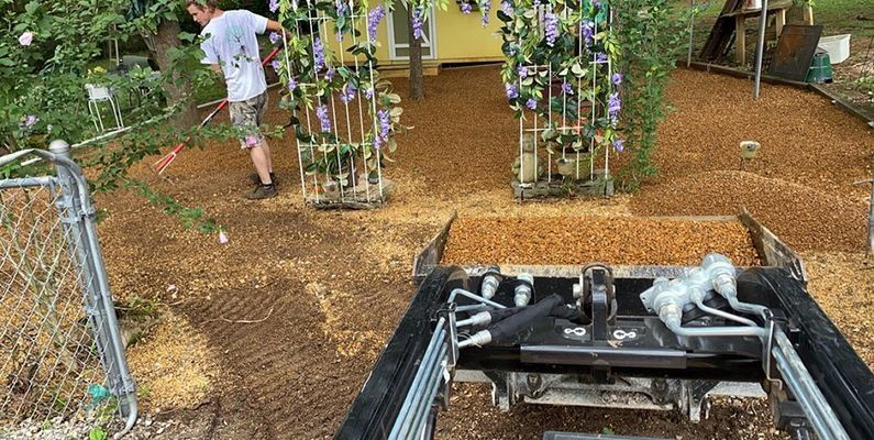 A person spreading mulch in a yard with a small tractor.  Arbor with purple flowers in background.