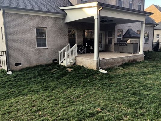 A light brick house with a covered patio, steps, and overgrown grass.
