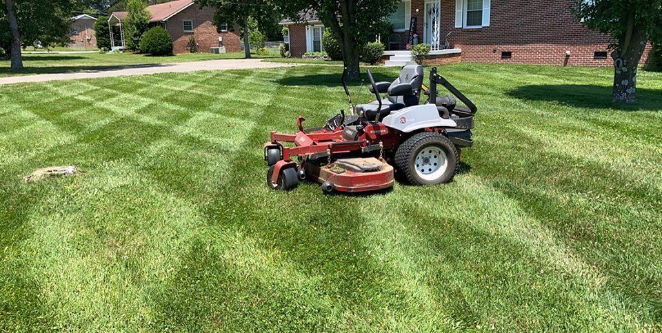 A person on a red and white lawnmower cutting grass in a diamond pattern in front of a house.