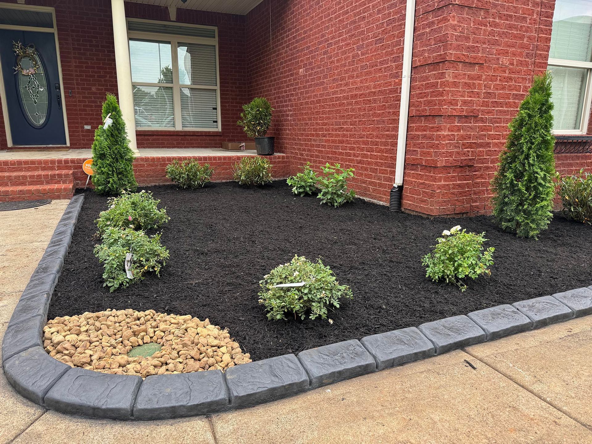 Landscaped front yard with black mulch, stone border, and small green bushes against red brick house.