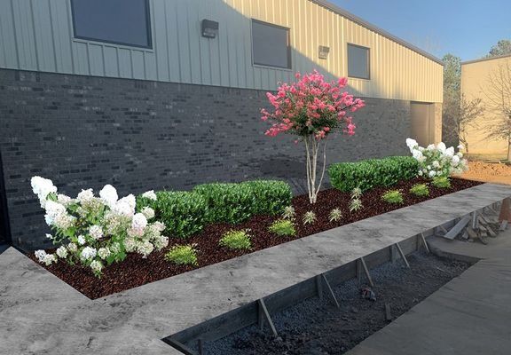 Flower bed with blooming pink tree, white hydrangeas, and green shrubs in front of a brick building.