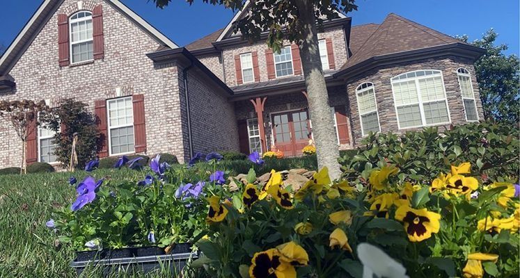 House with brick facade and red shutters, with flower bed of yellow and purple pansies in front.