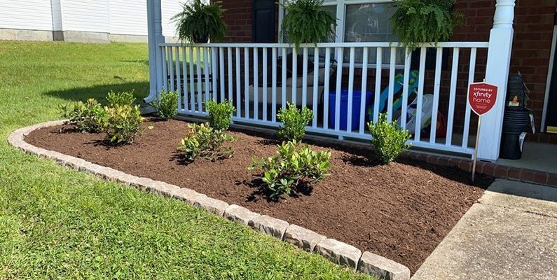 A well-maintained flower bed with small shrubs borders a porch with a white railing, brown mulch, and a grassy lawn.