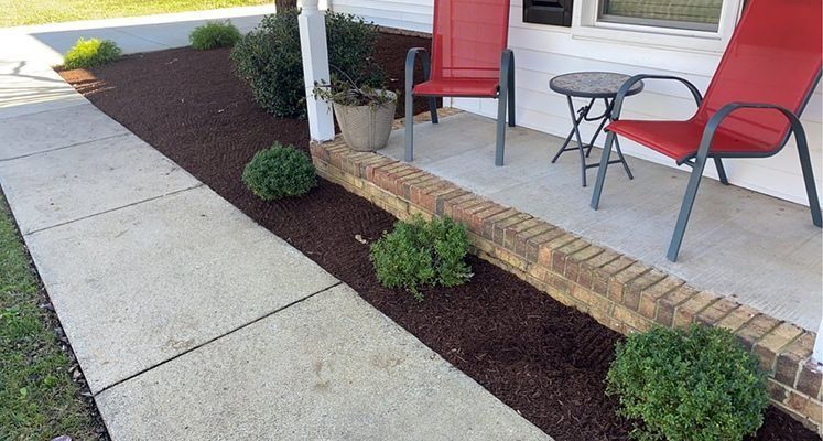 Front porch with two red chairs, small table, shrubbery, and walkway lined with dark mulch.