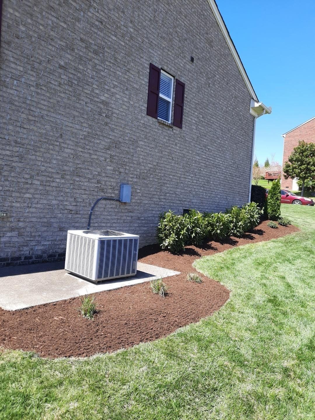 Air conditioner unit next to a brick building, with shrubs, mulch, and green grass.