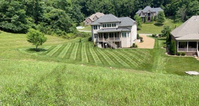 Lawn with striped patterns in front of a two-story house with a wrap-around porch. Green trees and grass.