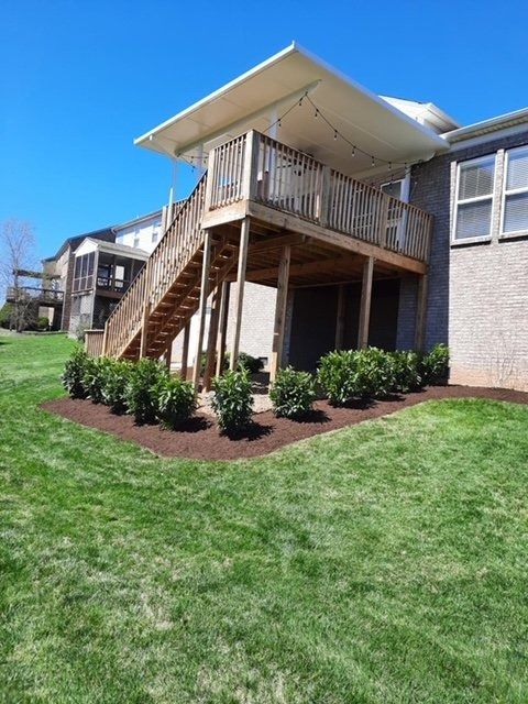 A multi-level wooden deck on a house with green grass, shrubs, and a clear blue sky.