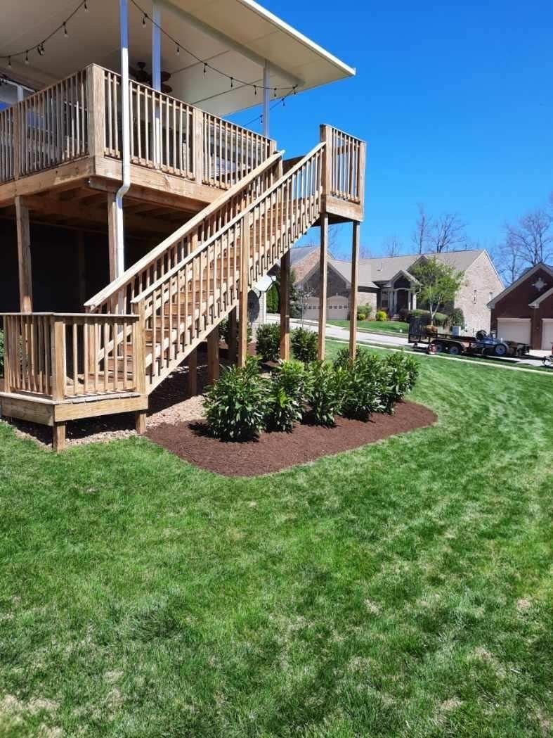 Wooden deck with stairs leading to a grassy yard, bushes with brown mulch, and a blue sky.