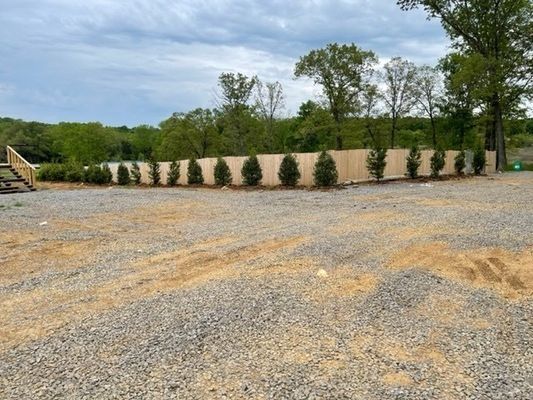 Gravel lot in front of wood fence with line of evergreen trees. Green trees and cloudy sky in background.