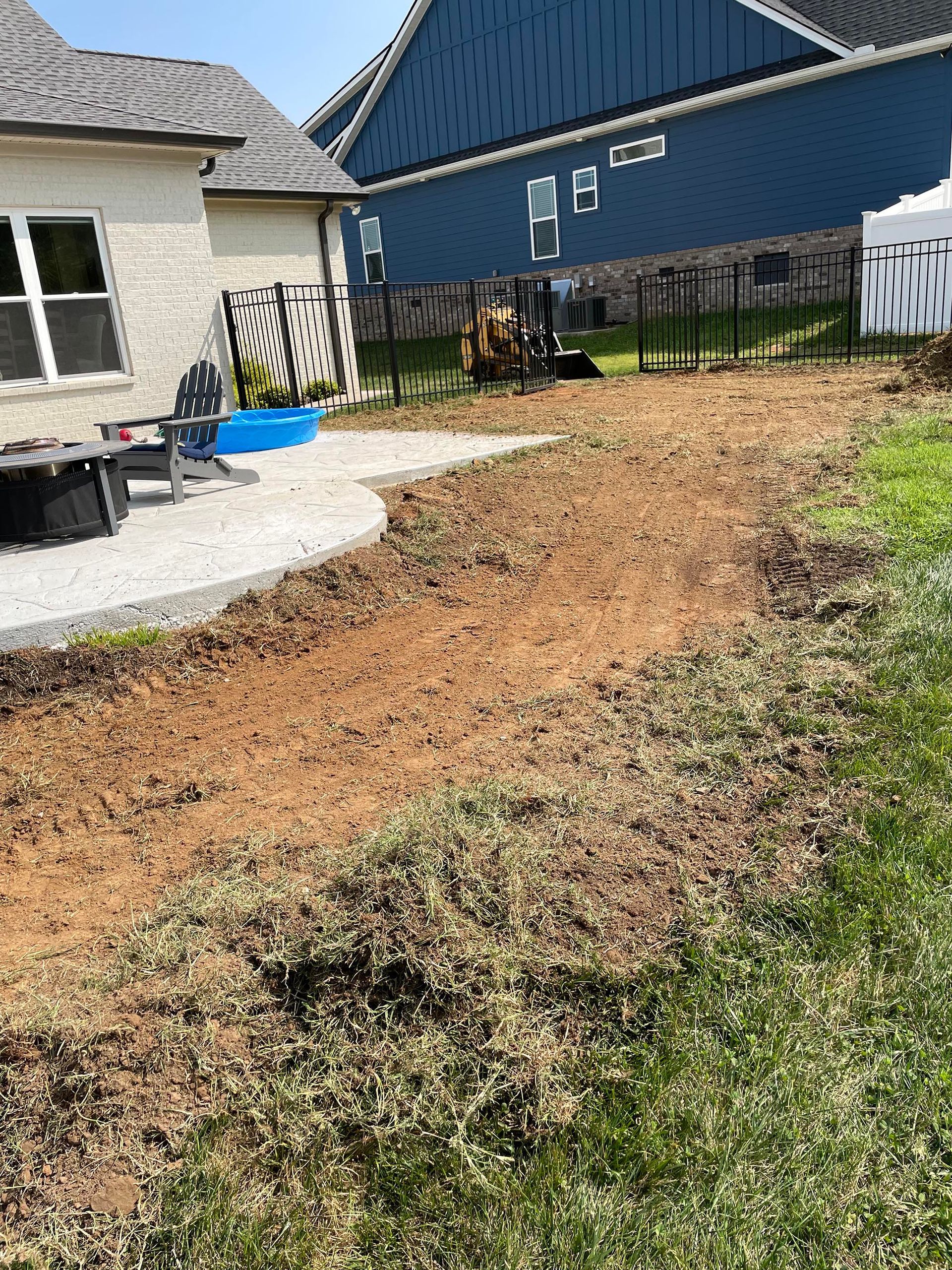 Dirt pathway leading to a fenced backyard, next to a stone patio. Grass and a blue house in the background.