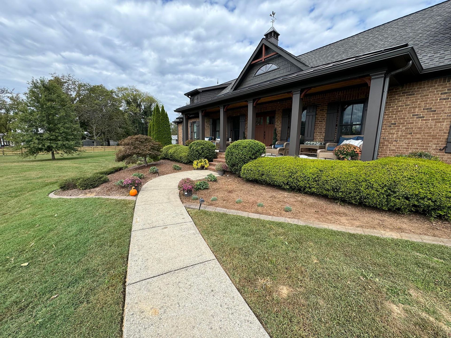 Stone path leads to a brick house with a covered porch, surrounded by greenery, under a cloudy sky.