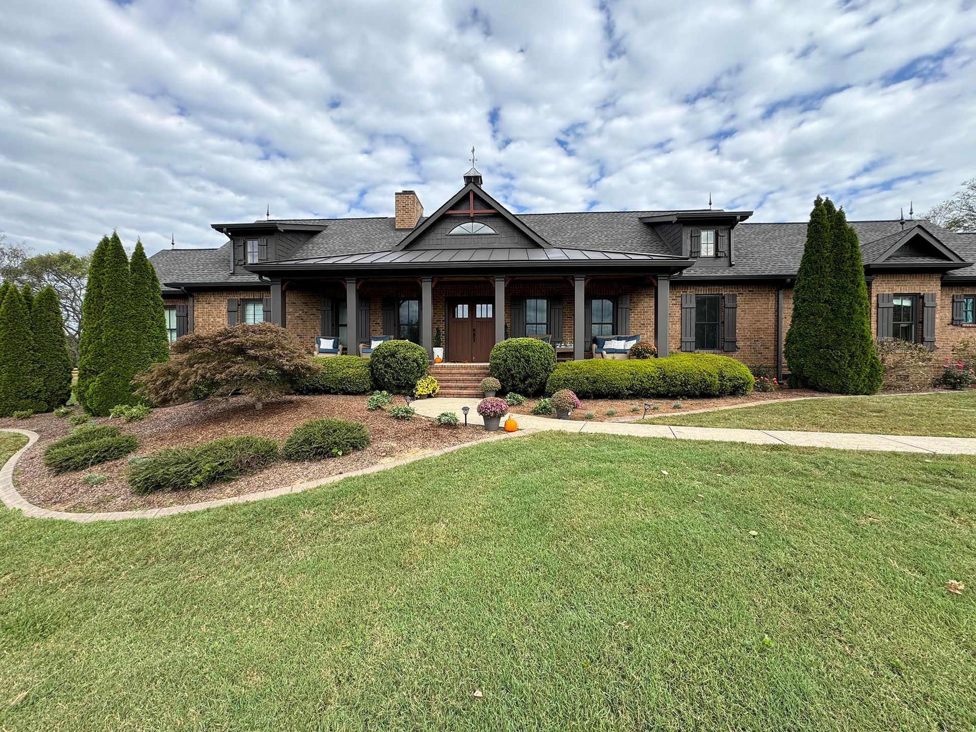 Brick house with a covered porch, brown roof, and landscaping under a cloudy sky.
