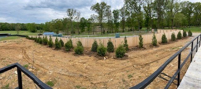 View of a newly planted landscape. Dirt, green shrubs, wooden fence, and trees in the background.