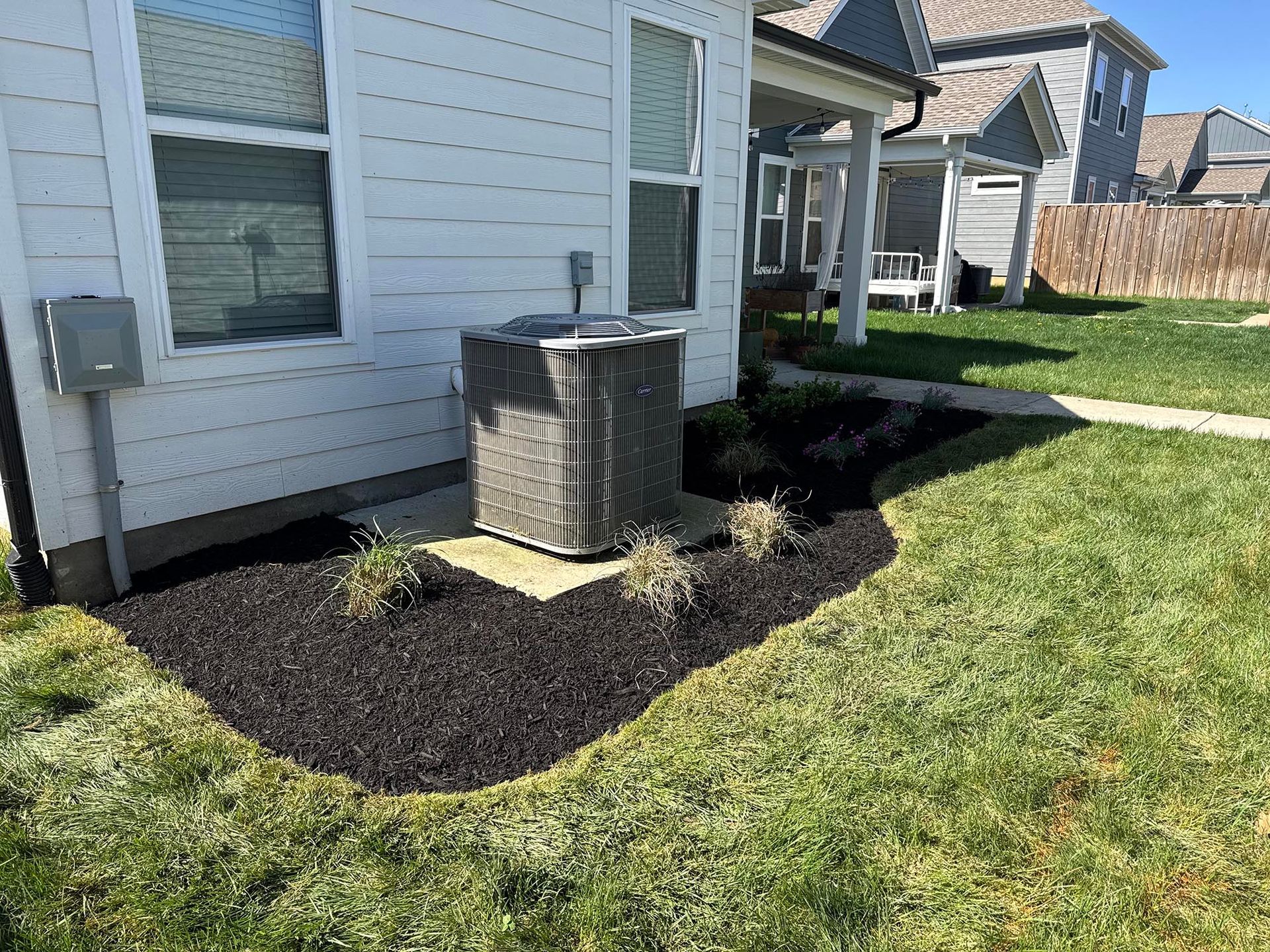 Air conditioner unit in a flower bed with black mulch next to a white house.