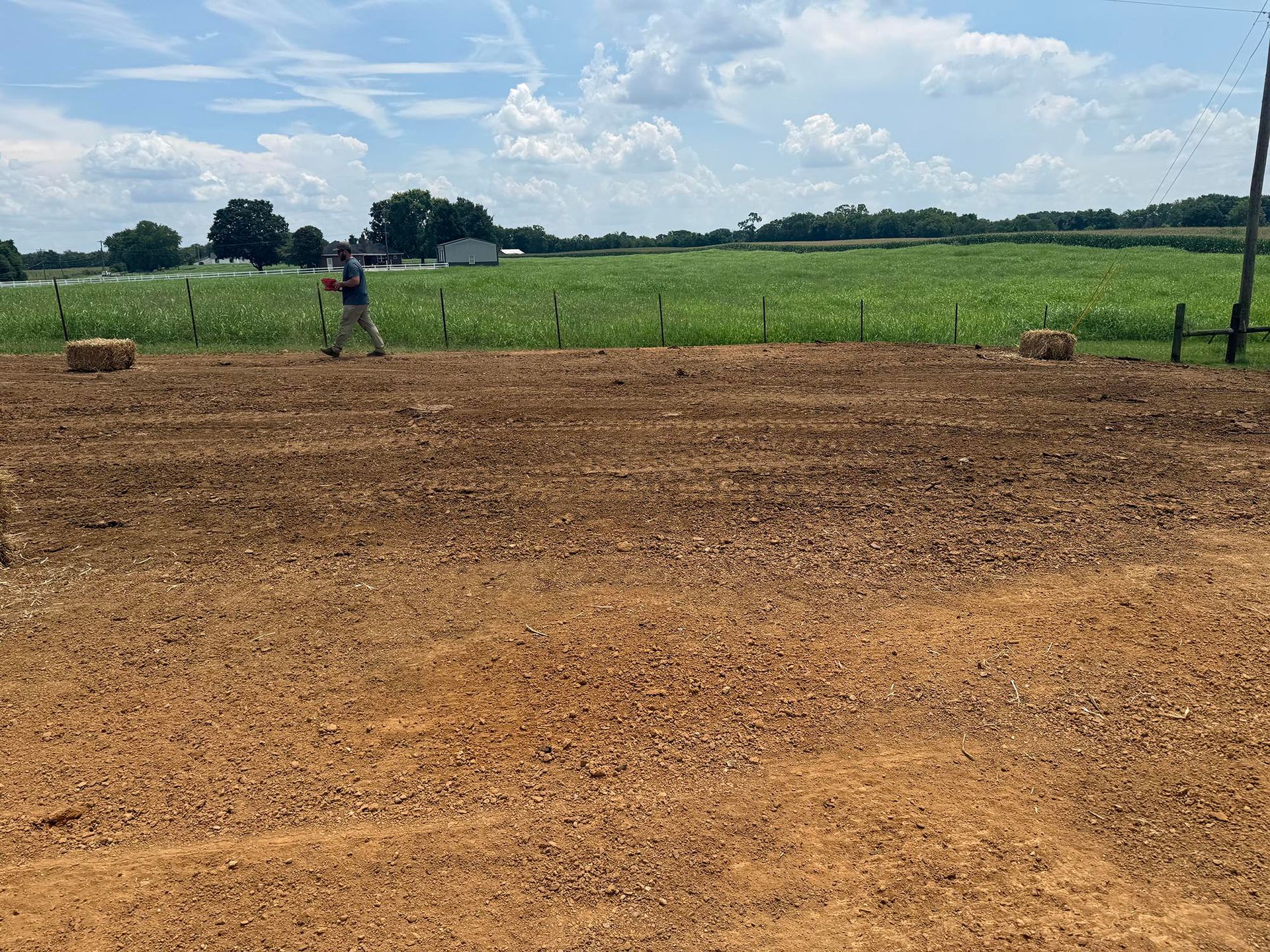 Person standing in a tilled field, next to hay bales, with a cornfield and sky in the background.