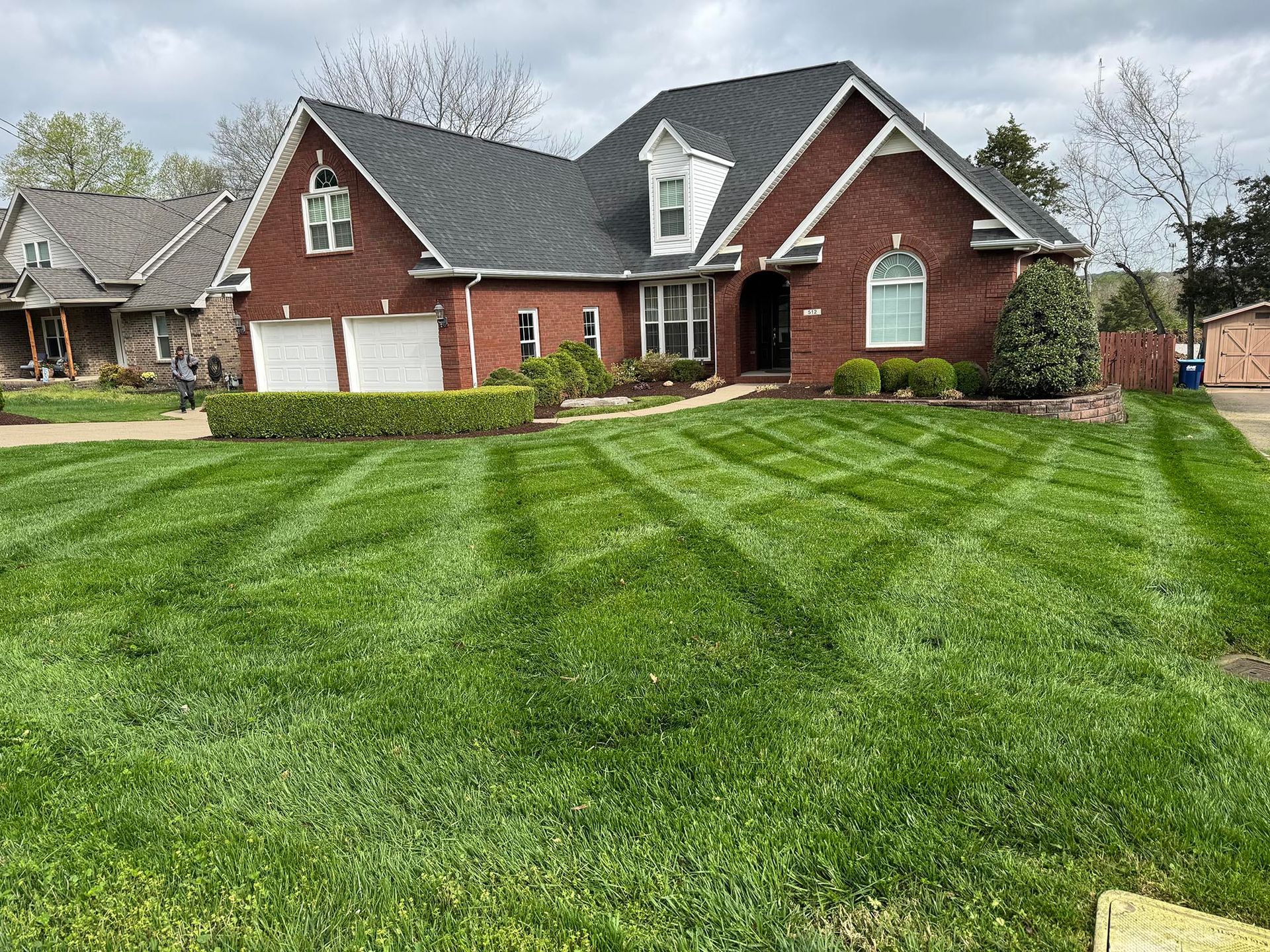 A brick house with patterned grass, hedges, and two garages. Overcast sky.