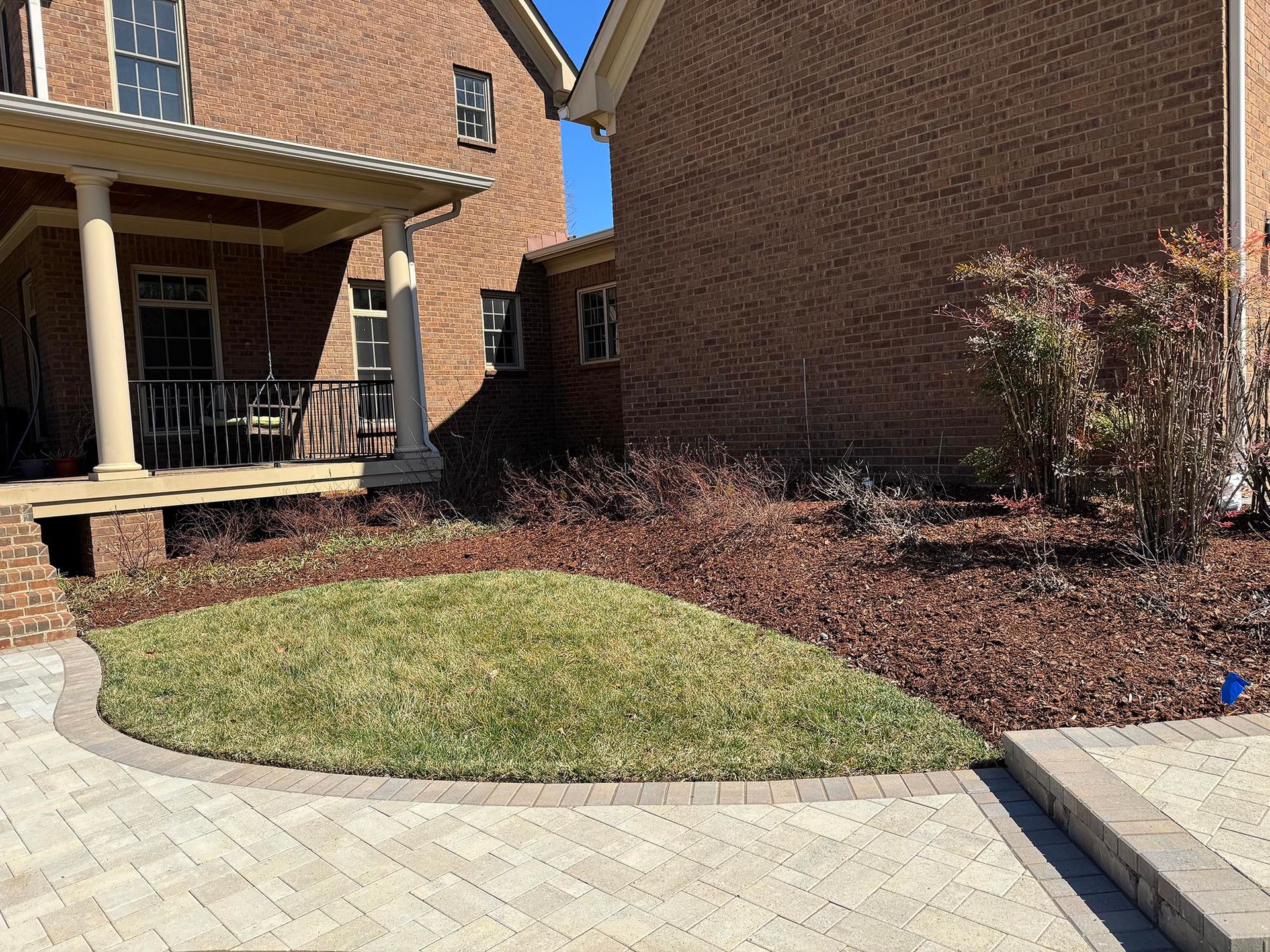A brick building with a porch, green lawn with brown mulch, and paved pathway on a sunny day.