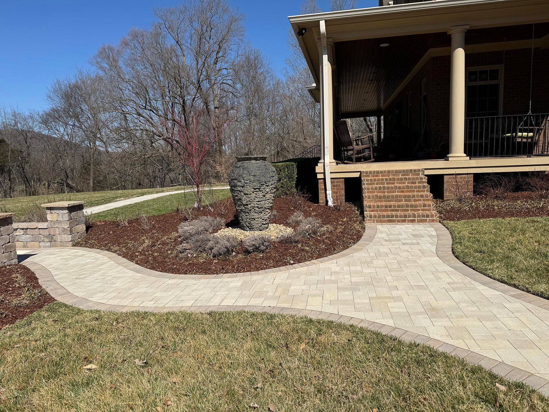 Stone pathway curves toward a house with a porch; a small fountain and landscaping are in between.