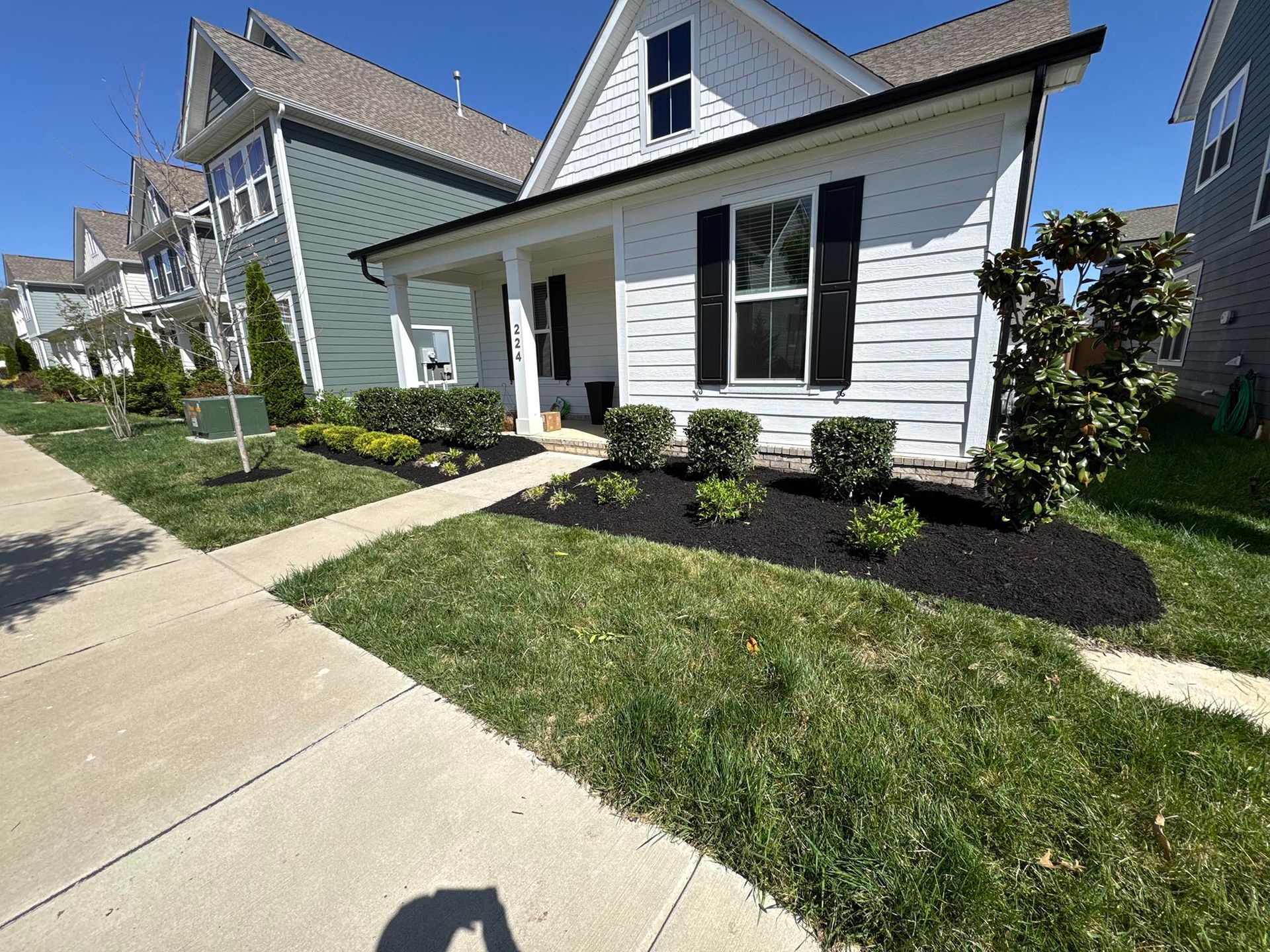 White house with black shutters, a small porch, and well-maintained landscaping, set in a neighborhood on a sunny day.