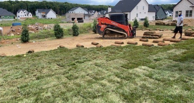 Construction site: worker laying sod. Orange skid steer, rolled sod, newly built houses in background.