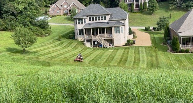 Lawn mower cutting grass on a sloped yard near a large house. Striped lawn pattern in progress.