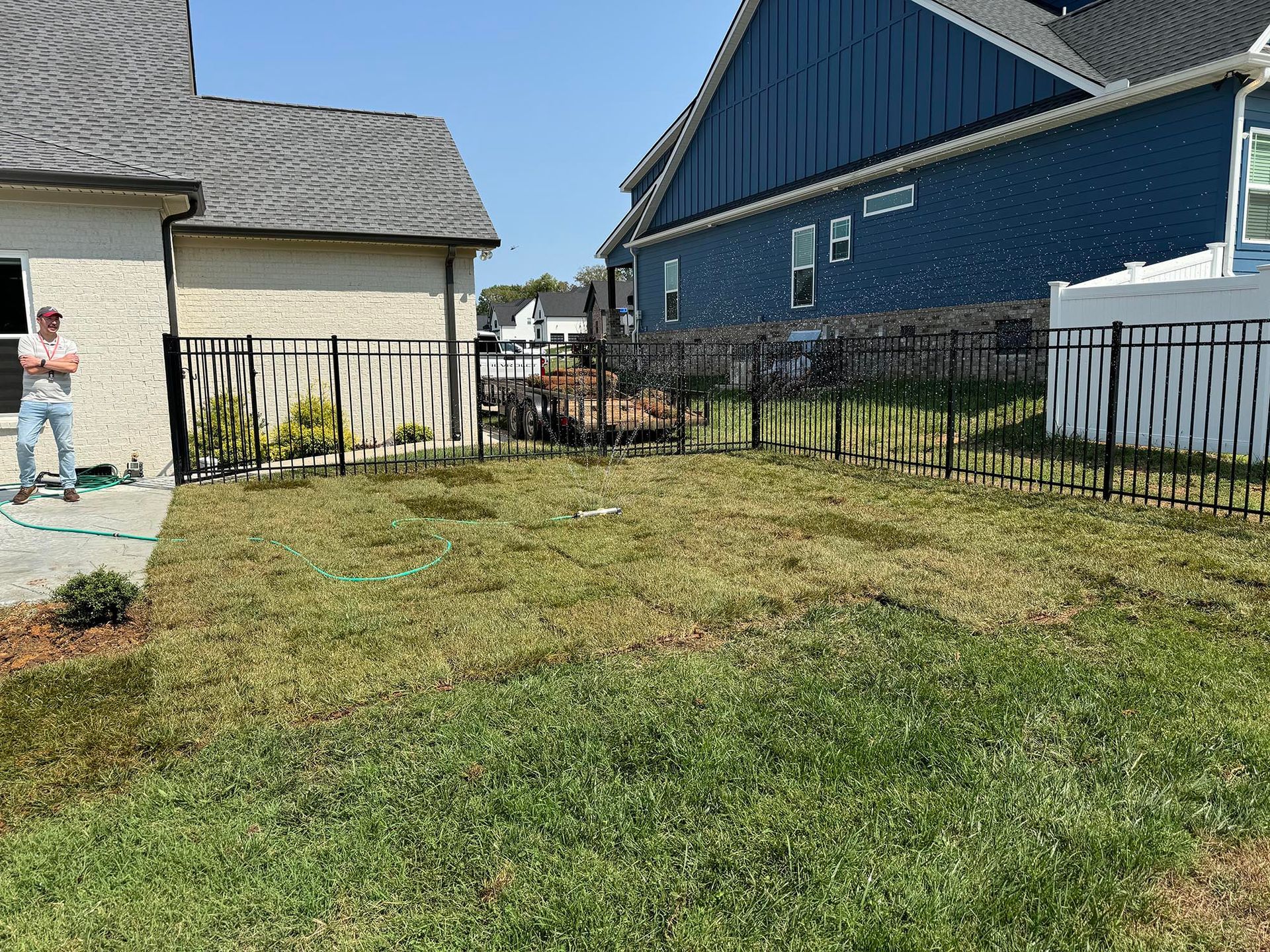 Man standing by a house with a patchy lawn, black fence, and blue house in the background on a sunny day.