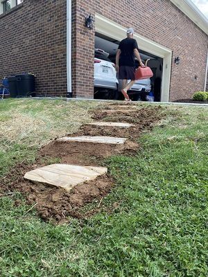 Person carrying a watering can walks up a path of stone steps in front of a garage.