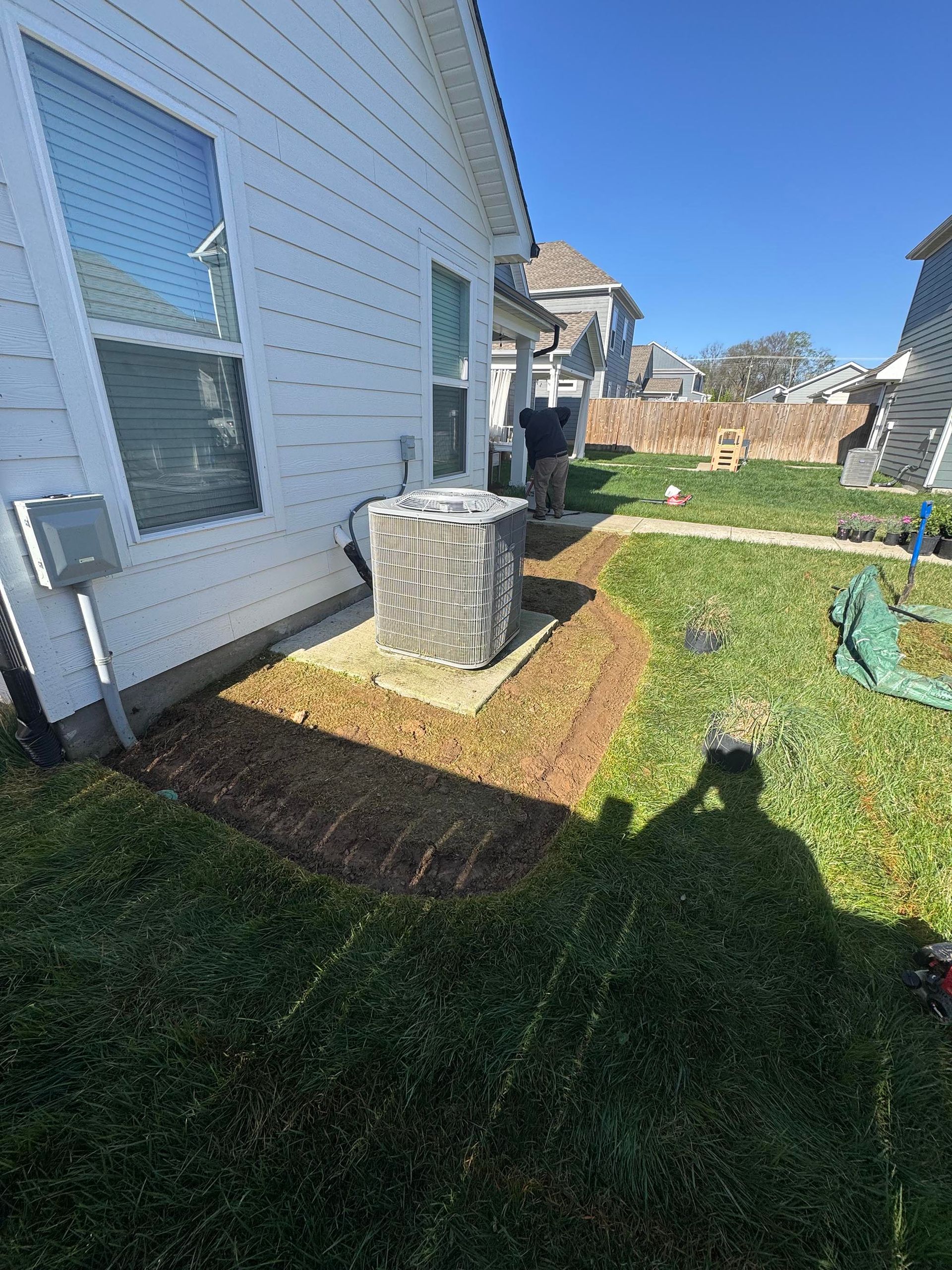 A person works on a landscaping project next to a house with an AC unit. Green grass and brown dirt are present.