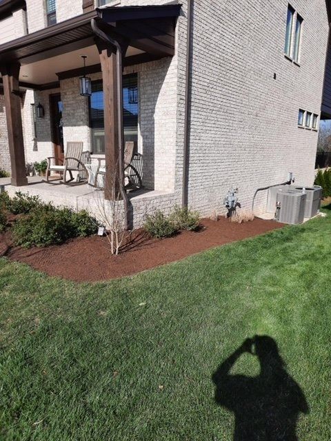 A house exterior with a porch, lawn, and mulch bed. A shadow of a person is cast on the grass.